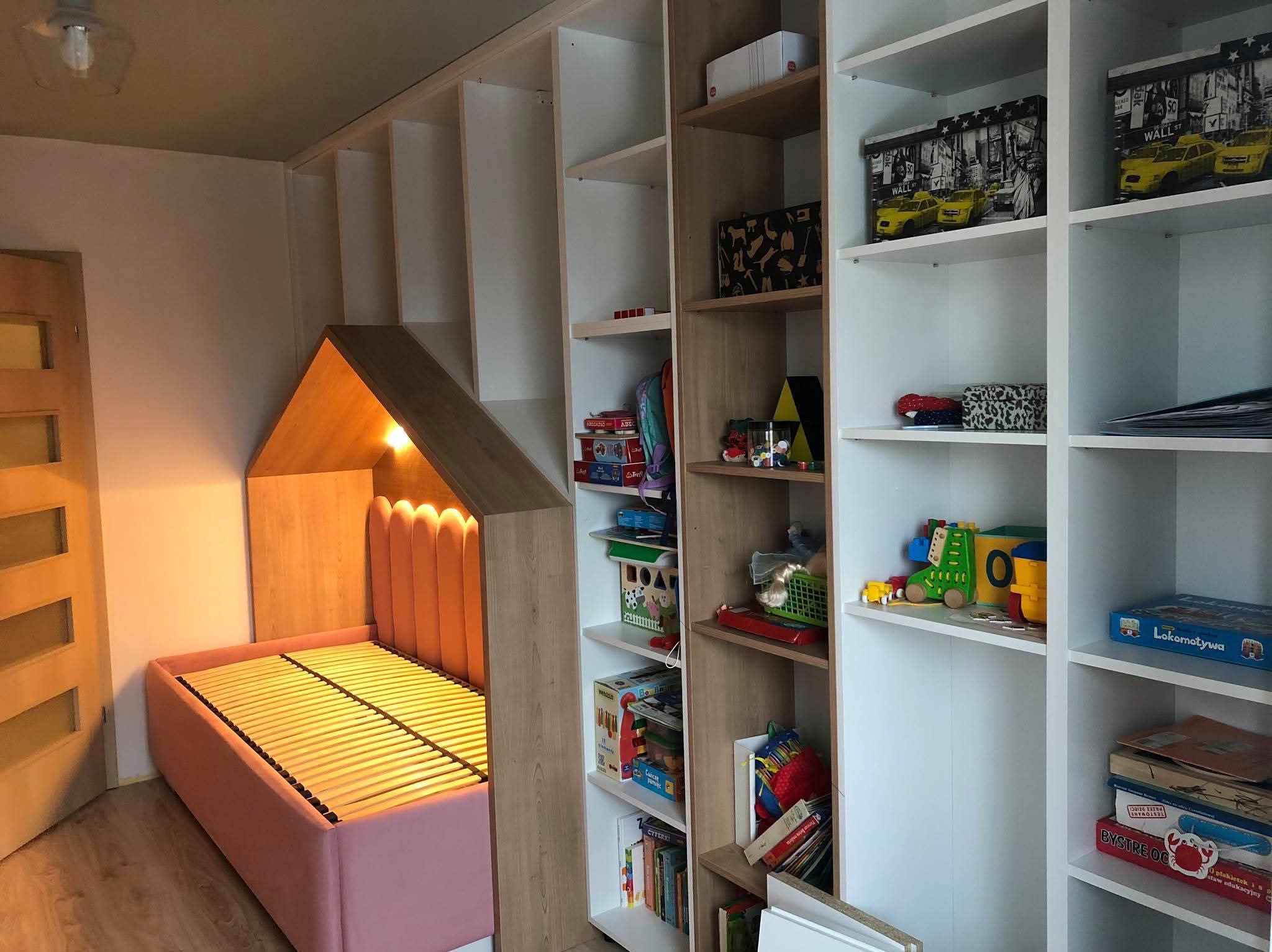 Pokój dziecięcy z zabudową meblową: różowe łóżko w kształcie domku z oświetleniem wewnętrznym, połączone z regałem na zabawki i książki, całość w odcieniach bieli, beżu i różu.