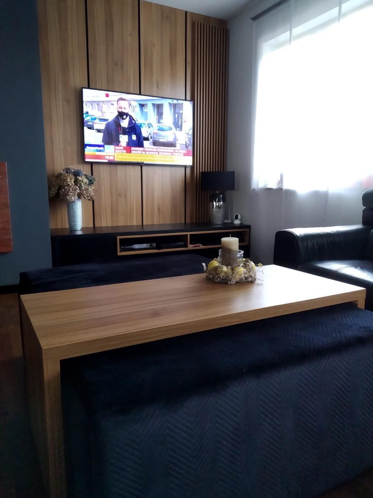 Nowoczesny salon z drewnianą ścianą telewizyjną, czarną szafką RTV, drewnianym blatem kawowym na pufie i skórzanym fotelem przy oknie.