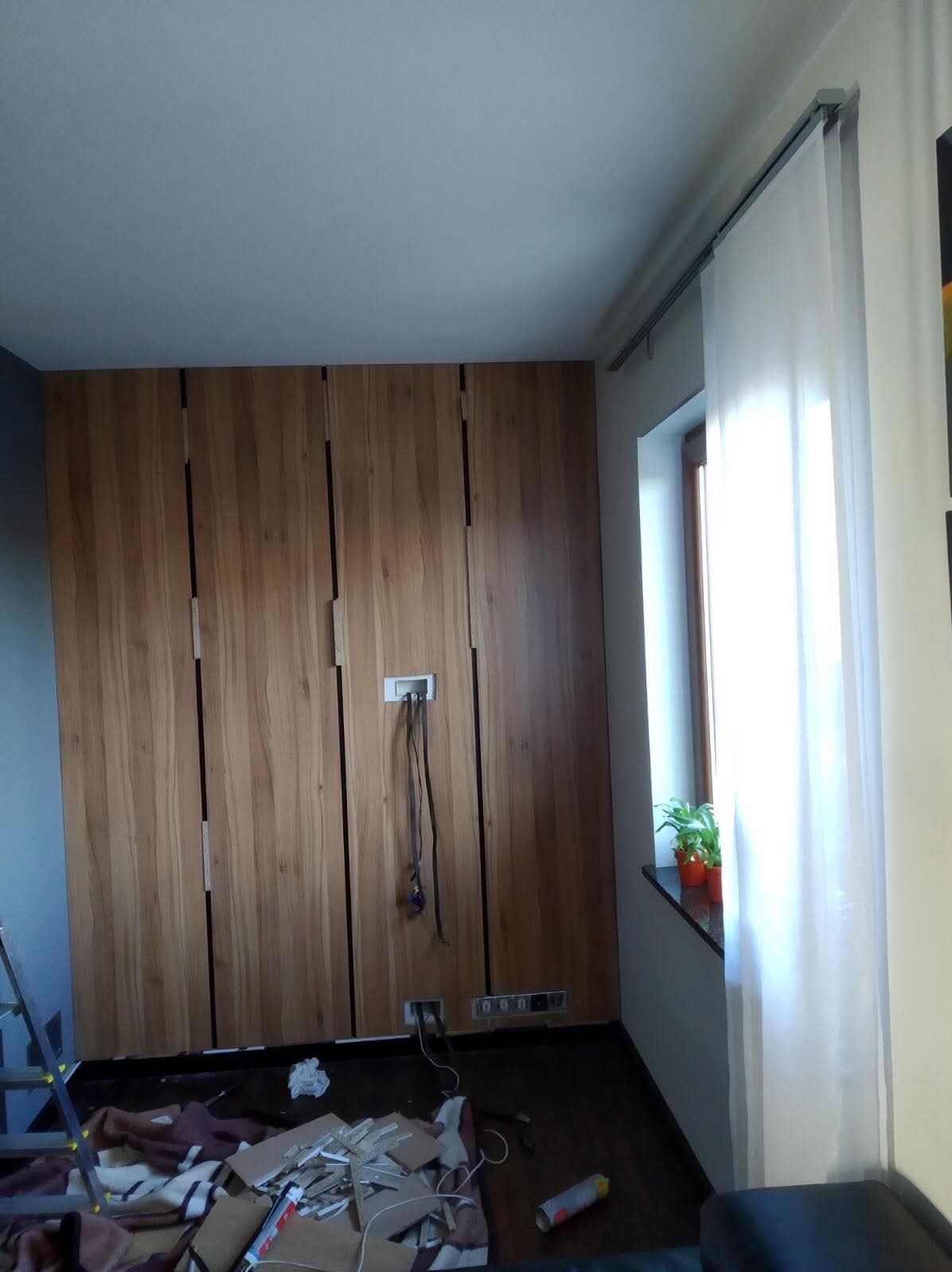 Montaż drewnianej zabudowy ściennej z ukrytymi drzwiami i instalacją elektryczną w trakcie prac wykończeniowych, widoczne kable, puszki i resztki materiałów na podłodze.