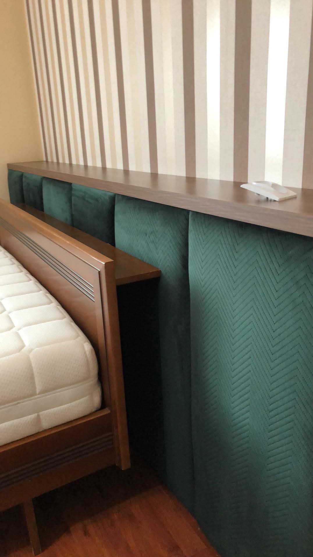 Tapicerowany zagłówek w kolorze butelkowej zieleni z geometrycznym wzorem, połączony z drewnianą półką i ramą łóżka z materacem, na tle ściany w pionowe pasy.