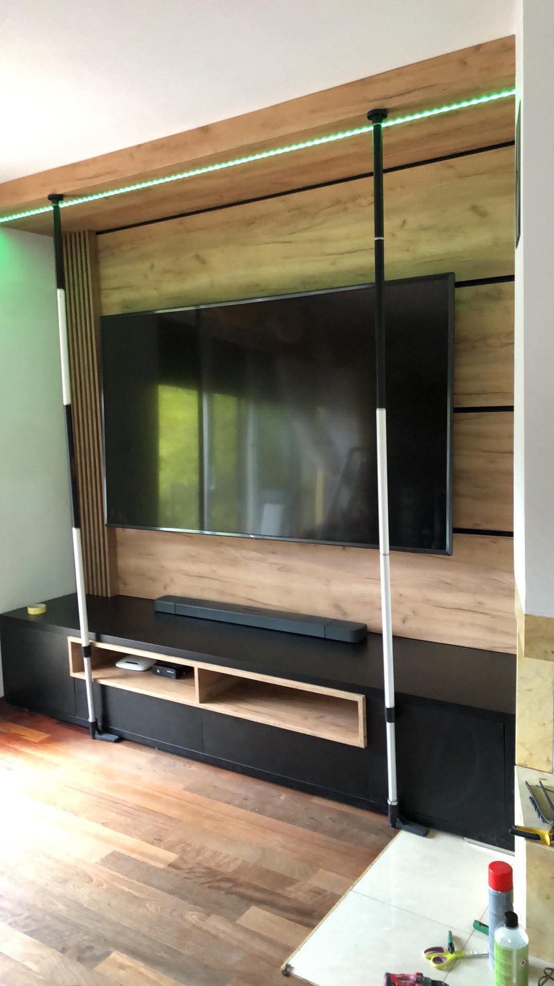 Nowoczesna ściana telewizyjna z drewnianą okładziną, podświetleniem LED, czarną szafką RTV i drewnianą podłogą, widoczne narzędzia na podłodze sugerują montaż w toku.