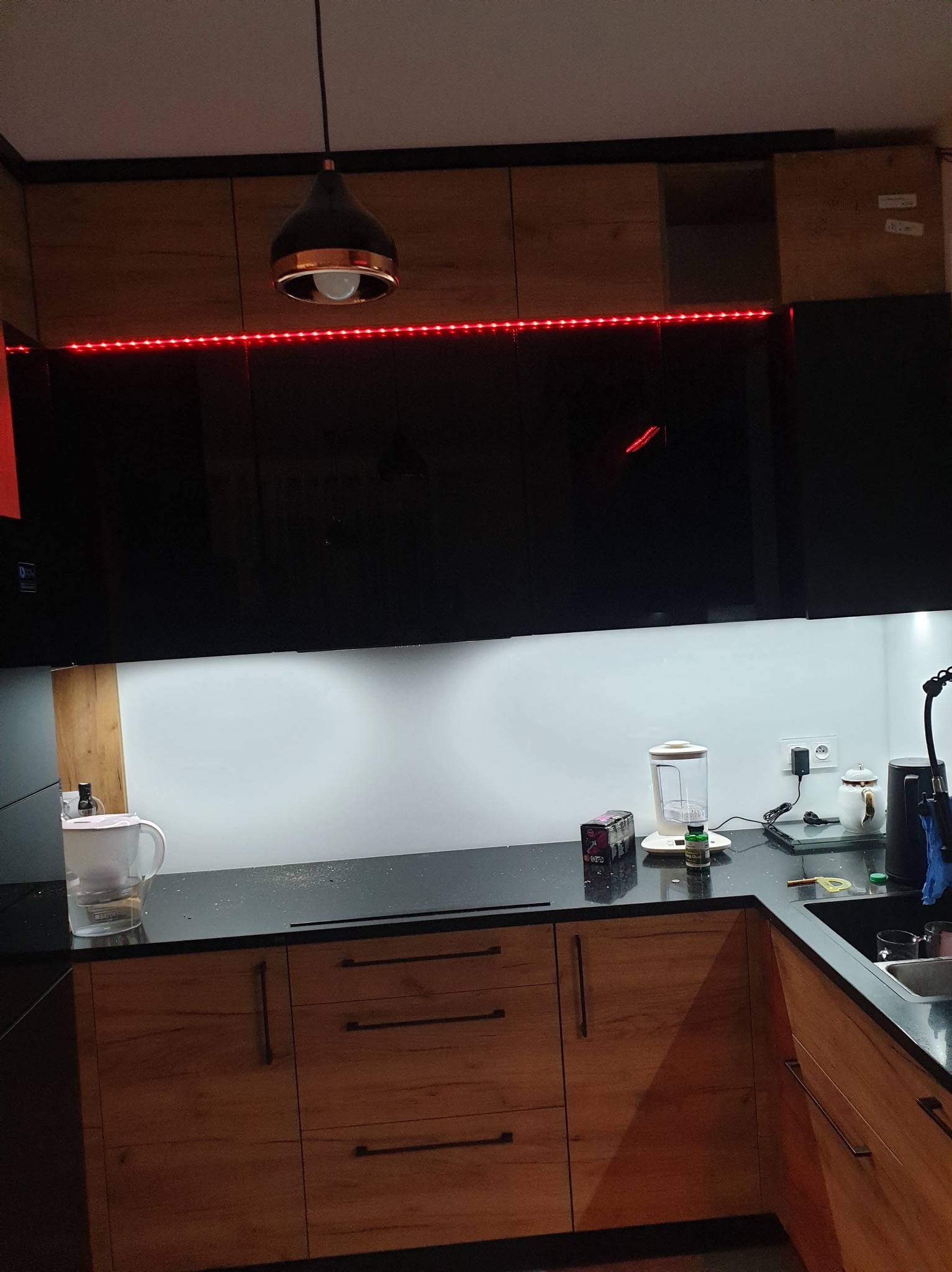 Nowoczesna kuchnia z drewnianymi frontami szafek dolnych i czarnymi szafkami górnymi, podświetlona czerwonymi ledami, z czarnym blatem i białym panelem ściennym.