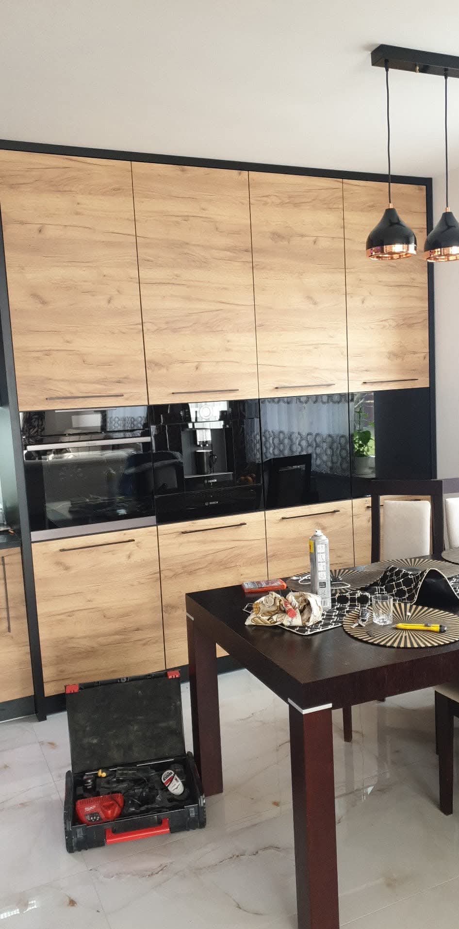 Nowoczesna kuchnia z drewnianymi frontami szafek wiszących i czarnymi, błyszczącymi szafkami dolnymi, widoczny otwarty kufer z narzędziami i akcesoriami na podłodze, stół z akcesoriami montażowymi.