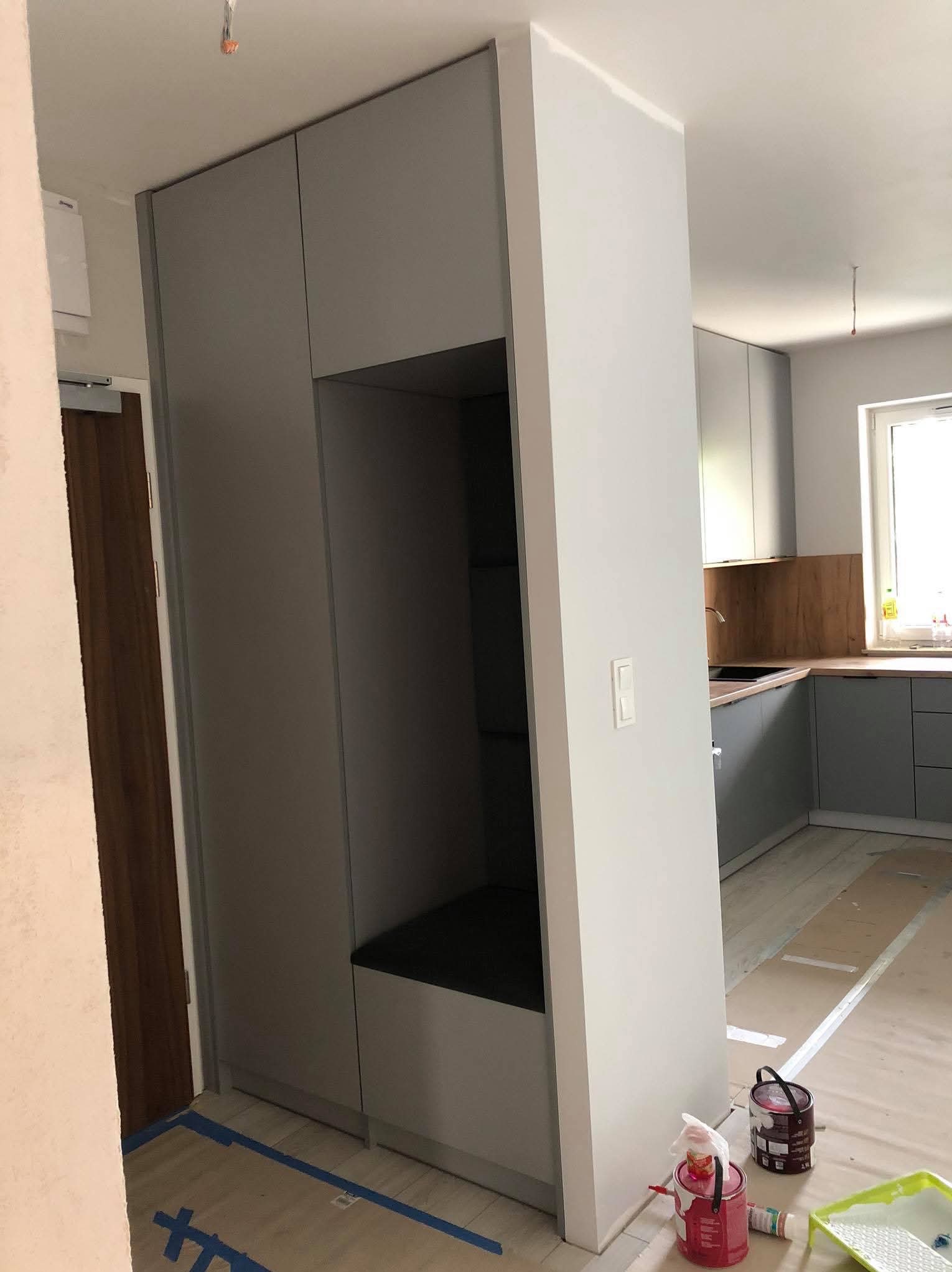 Szara zabudowa wnękowa z otwartą przestrzenią na siedzisko i półki, w tle widoczna kuchnia w podobnej kolorystyce, podłoga zabezpieczona taśmą malarską, puszki z farbą i akcesoria malarskie...