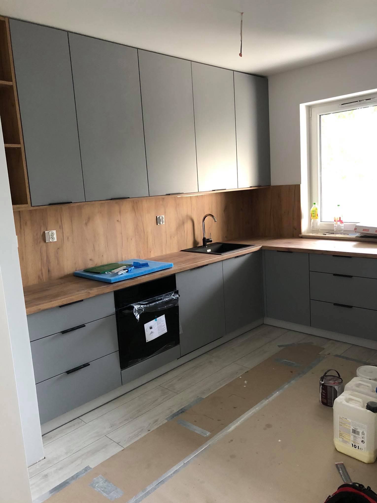 Szara zabudowa kuchenna z drewnianym blatem i panelem ściennym, czarny zlew i piekarnik. Widoczne zabezpieczenia podłogi podczas montażu.