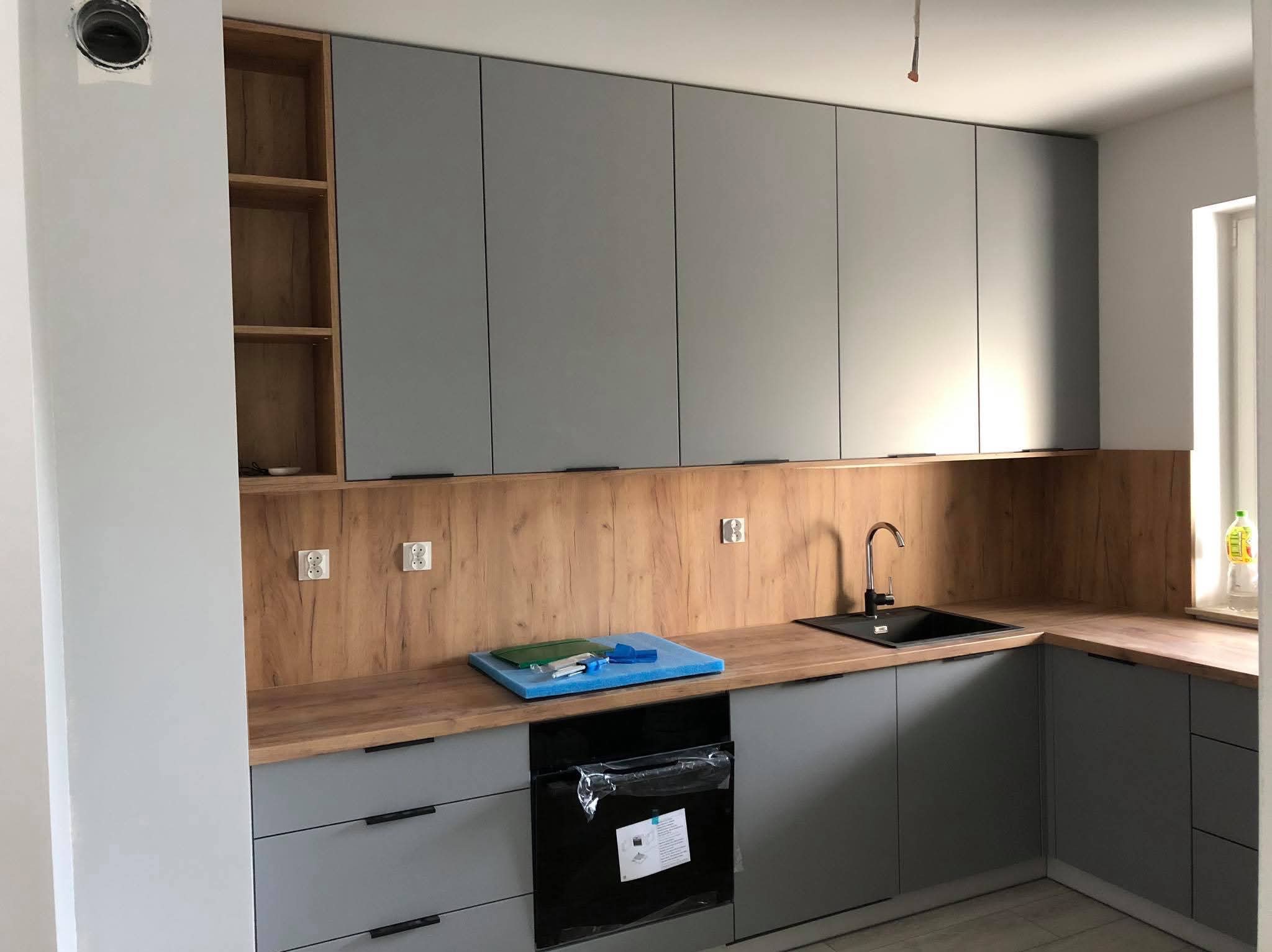 Szara kuchnia na wymiar z drewnianym blatem i okładziną, widoczne gniazdka elektryczne i akcesoria kuchenne na blacie.