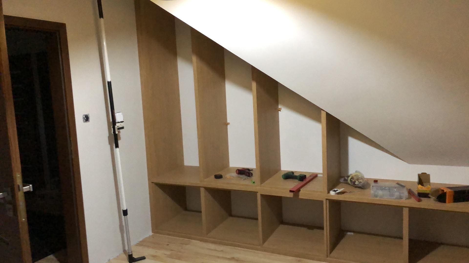 Montaż drewnianej zabudowy w skosie poddasza, widoczne narzędzia i materiały budowlane.