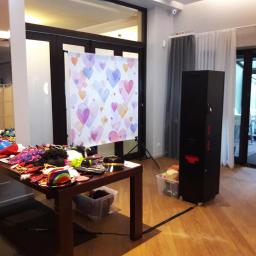 fotosbudka.pl - Stanowisko z fotobudką: stół z rekwizytami (kapelusze, peruki, okulary) przed tłem w serca, obok czarna fotobudka na kółkach, otwarte drzwi balkonowe w tle.