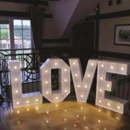 fotosbudka.pl - Świecący napis LOVE z dużych, białych liter, ustawiony na błyszczącej podłodze w eleganckim wnętrzu z elementami drewna, widok na okno w tle.