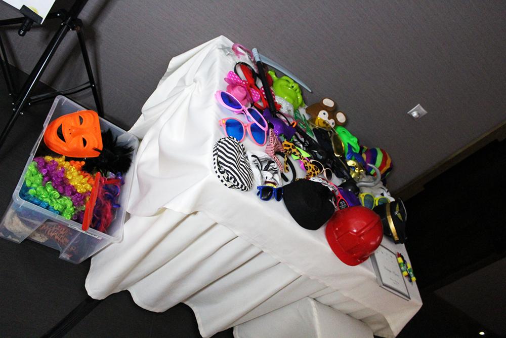 Stół przykryty białym obrusem z rekwizytami do zdjęć: kolorowe peruki, maski, okulary, kapelusze, plastikowa kosa, czerwony kask, umieszczone obok przezroczystego pojemnika z dodatkowymi akcesoriami.