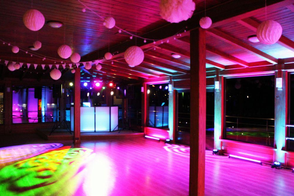 Drewniana sala eventowa z dekoracjami: papierowe kule, girlandy, oświetlenie sceniczne w odcieniach różu i błękitu, parkiet z kolorowymi projekcjami świetlnymi, stanowisko DJ-a w tle.