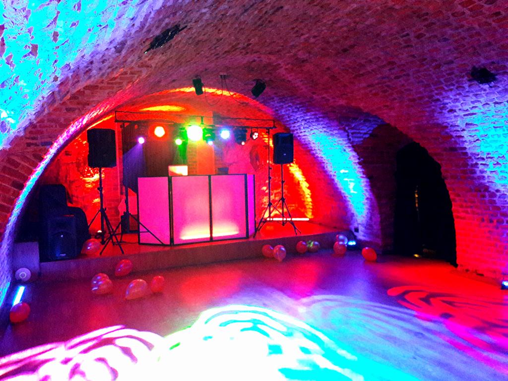 Scena z DJ-ką oświetloną kolorowymi światłami w ceglanym, sklepionym pomieszczeniu, z balonami rozrzuconymi na podłodze.