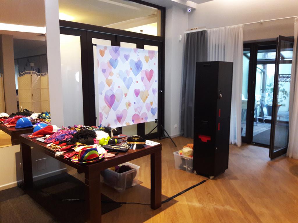 Stanowisko z fotobudką: stół z rekwizytami (kapelusze, peruki, okulary) przed tłem w serca, obok czarna fotobudka na kółkach, otwarte drzwi balkonowe w tle.