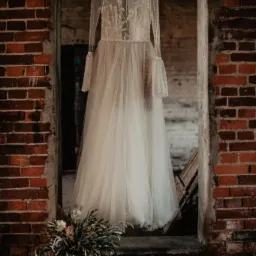 Delikatna suknia ślubna z długimi, koronkowymi rękawami, wisząca na wieszaku w starym, ceglanym przejściu. U dołu bukiet ślubny z zielonych gałązek i suszonych kwiatów.