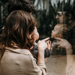 Pracownia KRA - Rodzina ogląda odbicie dziecka w szklanej tafli, matka wskazuje palcem na twarz dziecka, widoczne rośliny w tle.