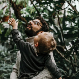 Pracownia KRA - Ojciec w okularach trzyma na rękach małe dziecko, oboje patrzą w górę na zwisające korzenie w otoczeniu zielonej roślinności w szklarni.