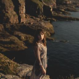 Pracownia KRA - Kobieta w zwiewnej sukience stoi na skalistym klifie z widokiem na morze, w tle strome, porośnięte trawą zbocza.