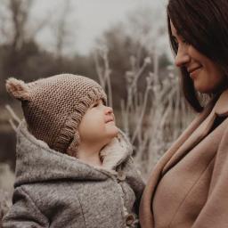 Pracownia KRA - Matka spogląda z uśmiechem na małe dziecko w wełnianej czapce z uszkami i szarym płaszczyku z futerkiem, na tle zimowej roślinności.