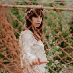 Pracownia KRA - Portret kobiety w koronkowej sukience i wianku na głowie, pozującej za pomarańczową siatką na tle zieleni.