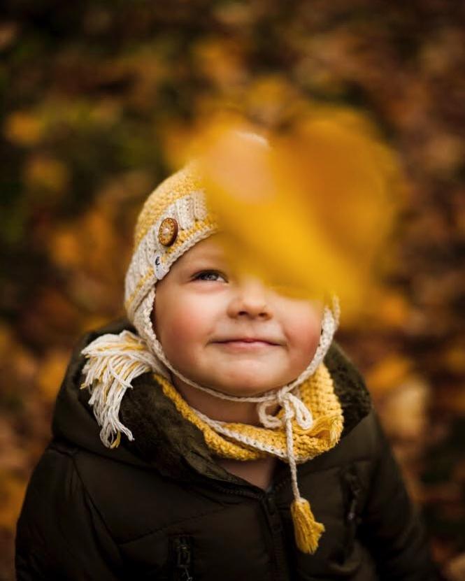 Uśmiechnięty chłopiec w żółtej czapce i szaliku, z jesiennym liściem przesłaniającym część twarzy, na tle rozmytych brązowych liści.