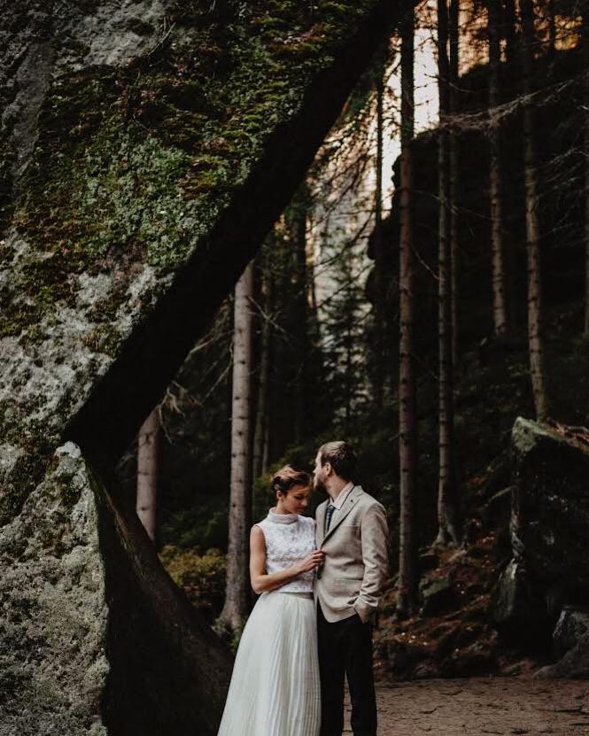 Para młoda w leśnej scenerii, panna młoda w białej sukni i koronkowej bluzce, pan młody w beżowej marynarce, stoją przy skalnej formacji porośniętej mchem, wysokie drzewa w tle.