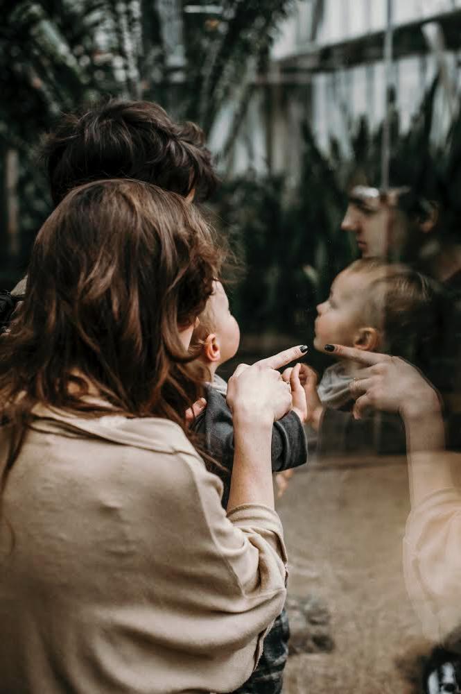 Rodzina ogląda odbicie dziecka w szklanej tafli, matka wskazuje palcem na twarz dziecka, widoczne rośliny w tle.