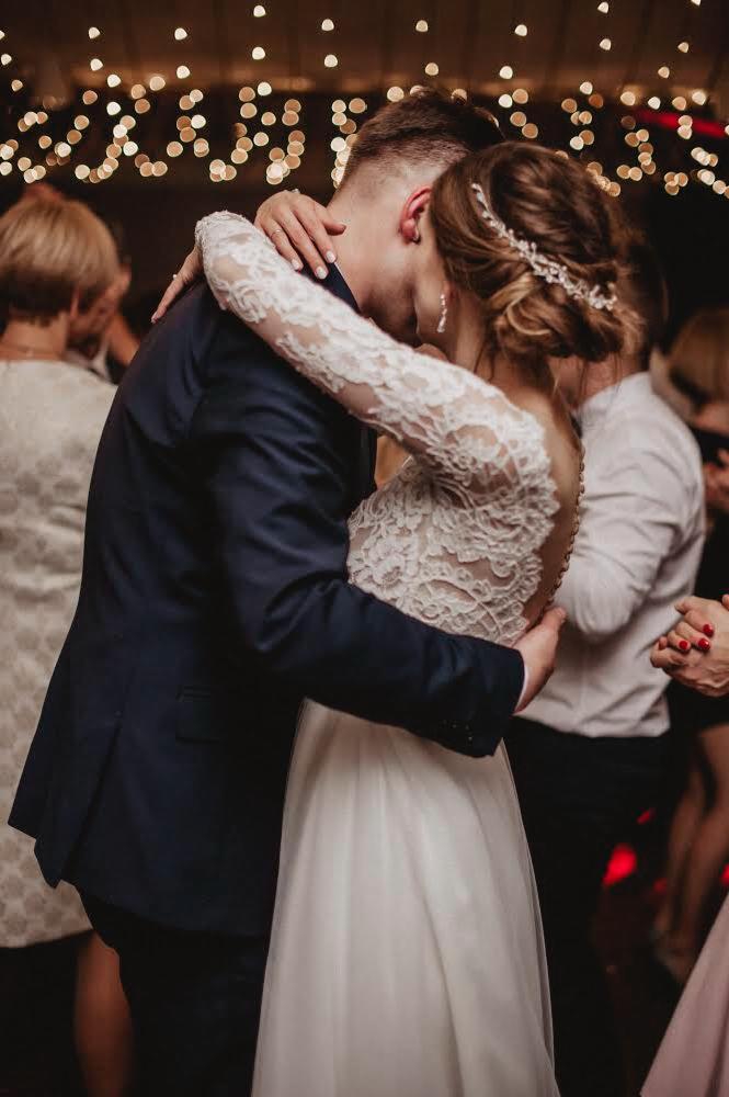 Para młoda w objęciach podczas pierwszego tańca, panna młoda w koronkowej sukni z długim rękawem i ozdobą we włosach, pan młody w granatowym garniturze, tło rozświetlone girlandami światełek.