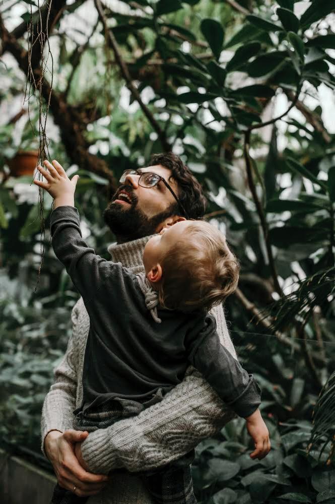Ojciec w okularach trzyma na rękach małe dziecko, oboje patrzą w górę na zwisające korzenie w otoczeniu zielonej roślinności w szklarni.