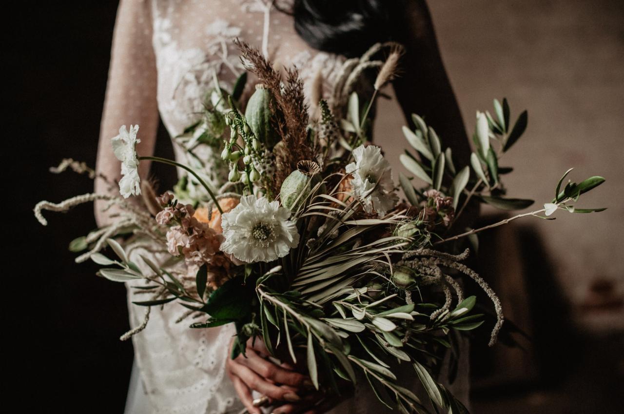 Panna młoda w koronkowej sukni trzyma w dłoniach bogaty bukiet z białych kwiatów, palmowych liści, traw i makówek w stylu rustykalnym.