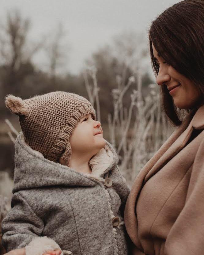 Matka spogląda z uśmiechem na małe dziecko w wełnianej czapce z uszkami i szarym płaszczyku z futerkiem, na tle zimowej roślinności.