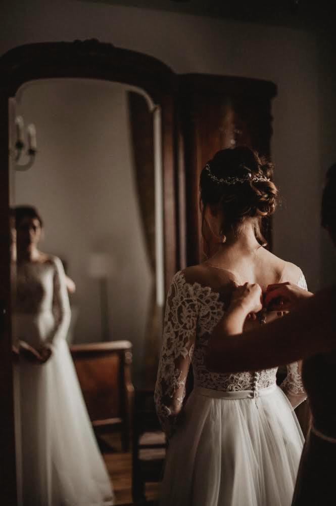 Panna młoda w sukni z koronkowymi rękawami, dopinana przez drugą osobę, widoczna w lustrze w tle.