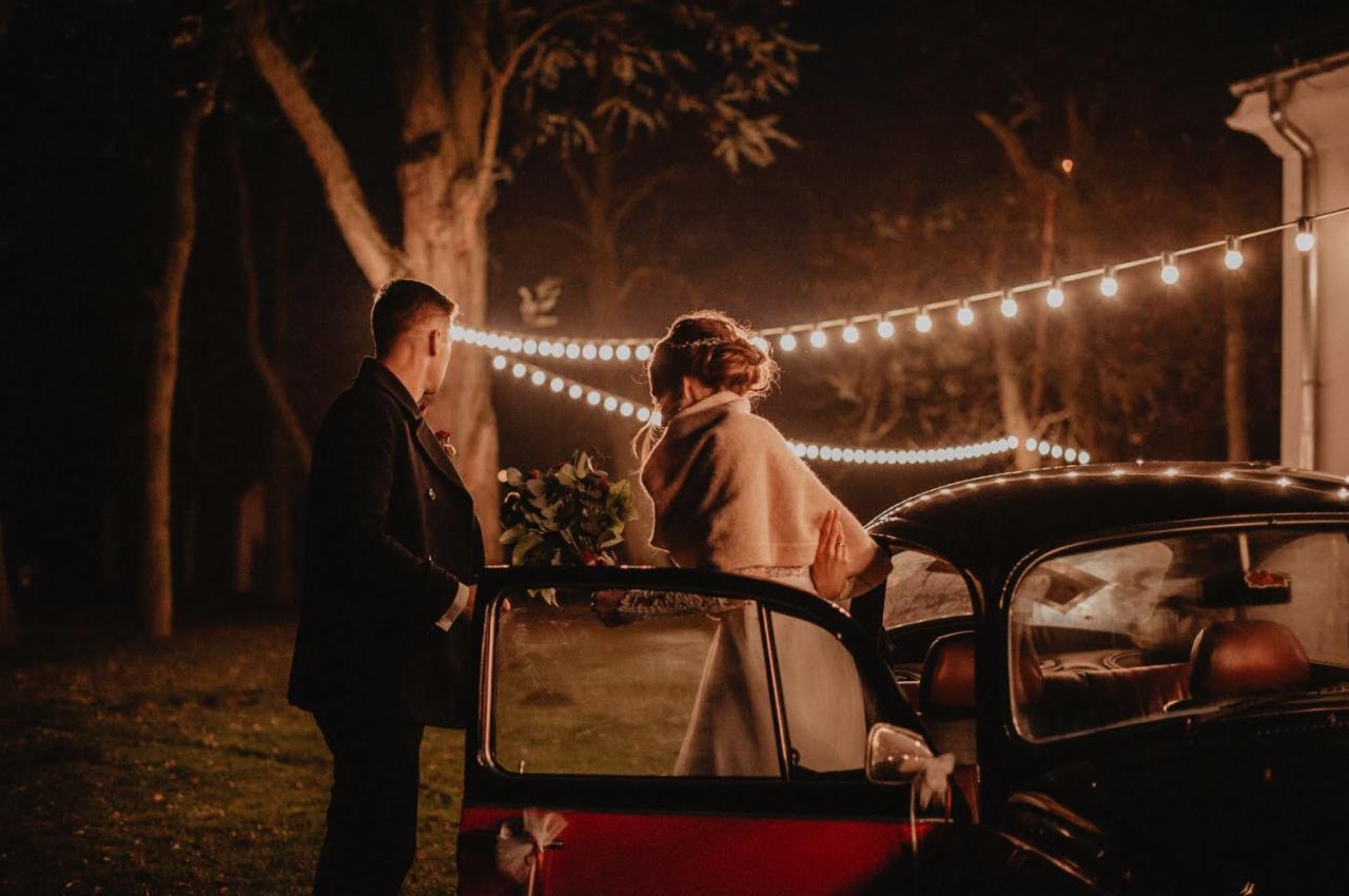 Para młoda przy czarnym, zabytkowym samochodzie w wieczornej scenerii, oświetlonej sznurem żarówek.
