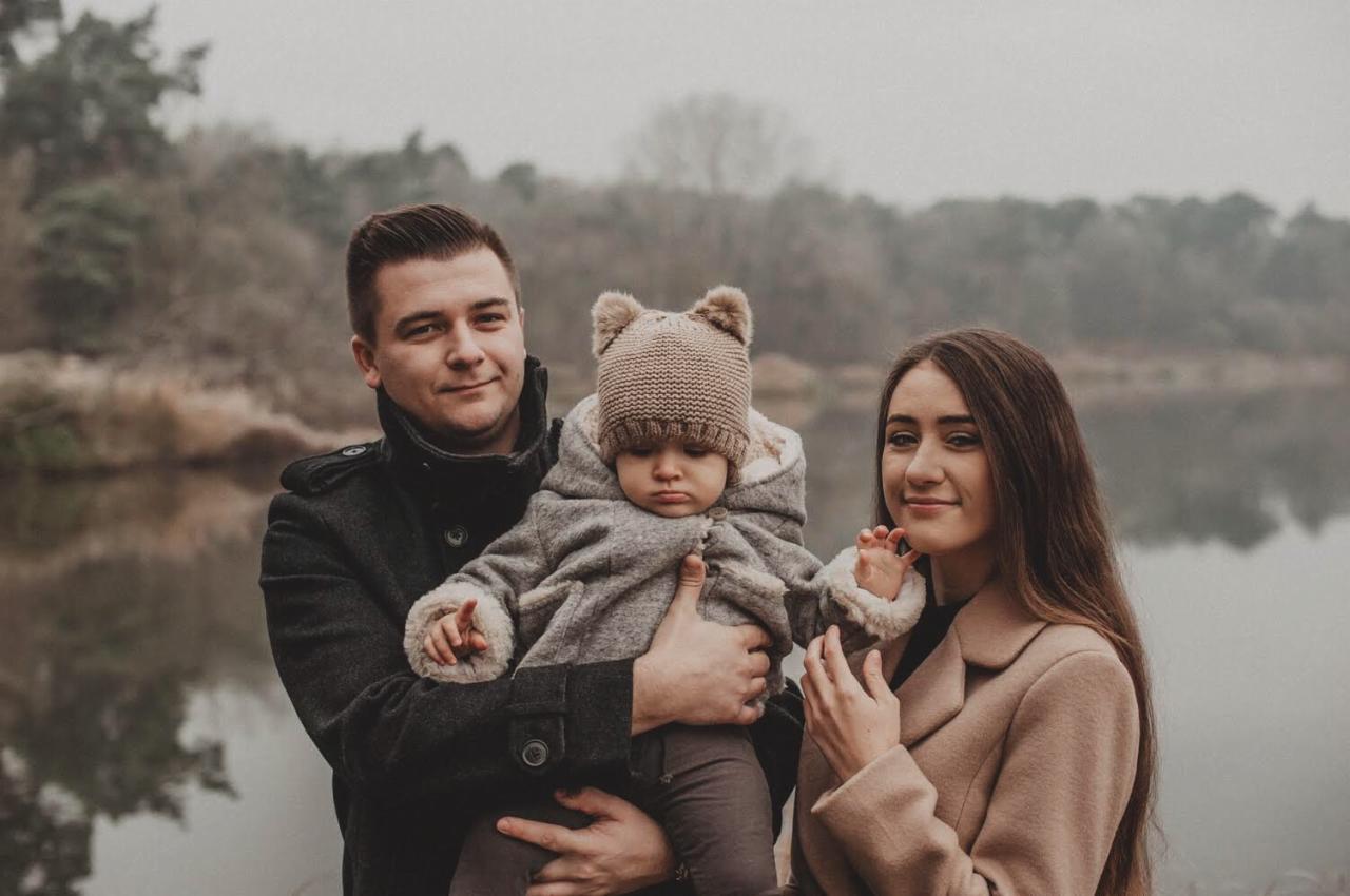 Rodzinne zdjęcie portretowe: ojciec trzyma dziecko w szarej kurtce z uszami, matka obok w beżowym płaszczu, na tle jeziora i drzew w pochmurny dzień.