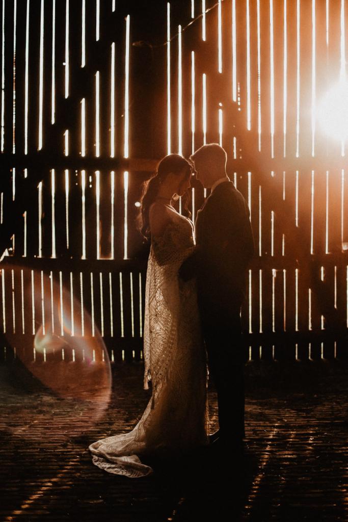 Para młoda w objęciach na tle drewnianej ściany z prześwitami, przez które wpada ciepłe światło, tworząc efektowne cienie na brukowanej podłodze.