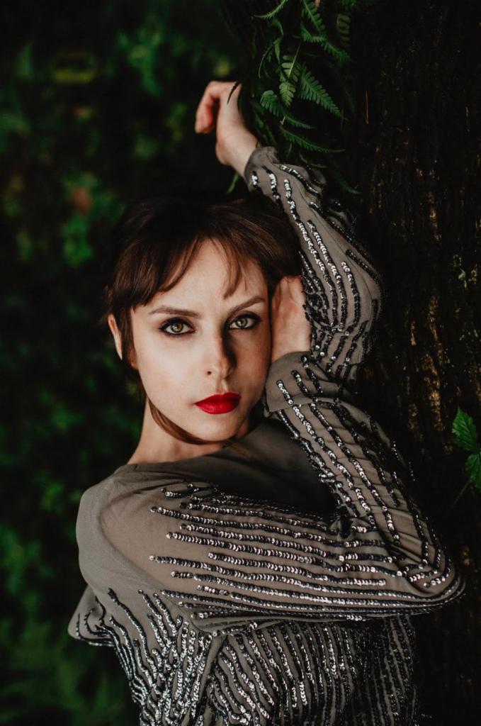 Portret kobiety z intensywnym makijażem, opierającej się o korę drzewa na tle zieleni, ubrana w szarą, zdobioną cekinami bluzkę.