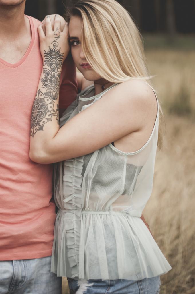 Portret pary w plenerze: blondwłosa kobieta opiera głowę o ramię mężczyzny w koralowej koszulce, eksponując tatuaż na ramieniu, tło stanowi wysoka trawa.