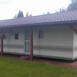 Parterowy, podłużny dom mobilny z brązowym dachem krytym dach&oacute;wką, drewnianymi słupami podtrzymującymi daszek wzdłuż elewacji i niewielkim tarasem z czerwonej kostki.