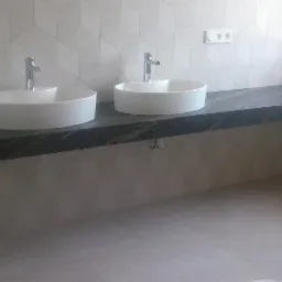 Dwie białe, okrągłe umywalki nablatowe z chromowanymi bateriami na blacie z ciemnego kamienia w łazience z geometryczną białą mozaiką na ścianie.