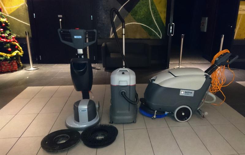 Profesjonalny sprzęt do czyszczenia podłóg w pomieszczeniu z choinką w tle: szorowarka jednotarczowa, odkurzacz przemysłowy i automat szorująco-zbierający marki Nilfisk.