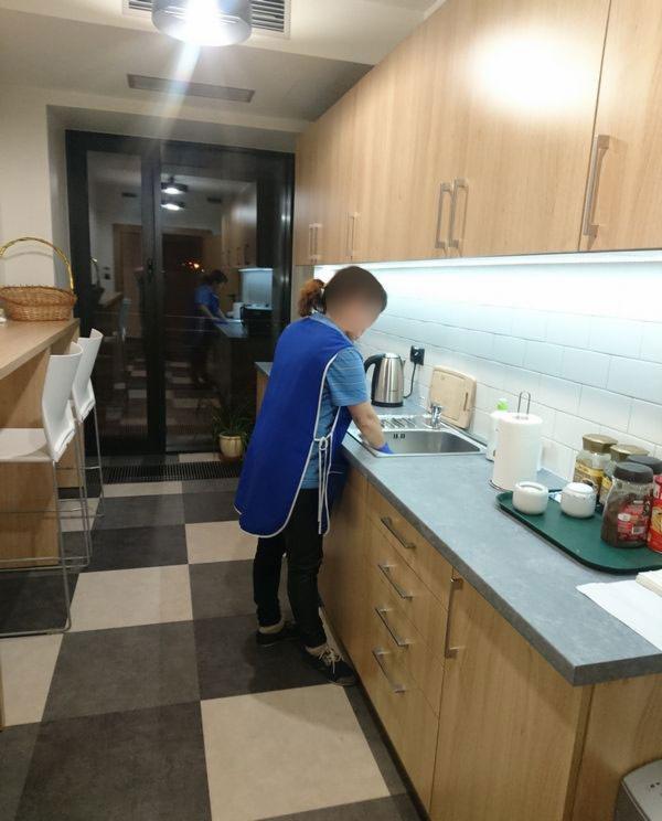 Osoba w niebieskim fartuchu, z założonymi rękawicami, myje zlew w kuchni z drewnianymi szafkami i białymi płytkami na ścianie.