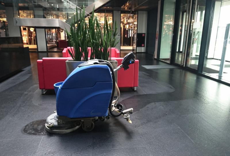 Automat szorująco-zbierający w trakcie pracy na ciemnej, błyszczącej posadzce w holu biurowca, widoczne czerwone sofy i zieleń w donicach.