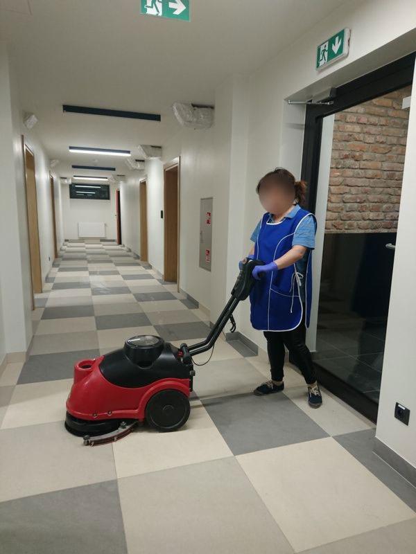 Pracownica w niebieskim fartuchu i rękawicach obsługuje czerwoną maszynę do czyszczenia podłóg w długim korytarzu z szachownicową posadzką.