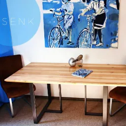 Jasny drewniany blat biurka na metalowych nogach w minimalistycznym stylu, z dekoracjami i obrazem z motywem rowerowym w tle.