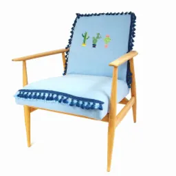Jasnoniebieski fotel z drewnianymi ramionami, zdobiony ciemnoniebieskimi frędzlami i haftowanymi kaktusami na oparciu.