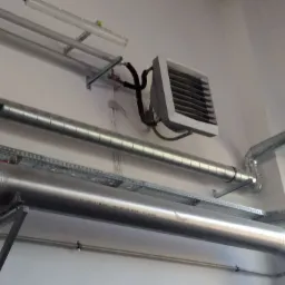 Szare rury wentylacyjne zamocowane do ściany, z widocznym grzejnikiem przemysłowym z czarnymi przewodami i oświetleniem sufitowym w tle.