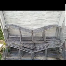 Szkielet z prętów zbrojeniowych ułożony w kształt przypominający domki, na tle betonowej ściany i fragmentu zieleni.