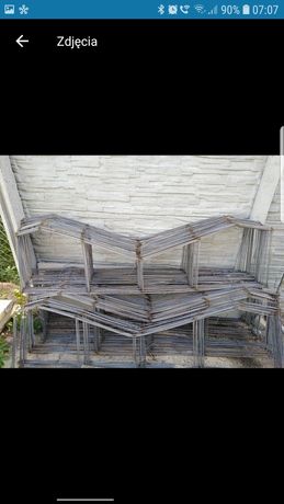 Szkielet z prętów zbrojeniowych ułożony w kształt przypominający domki, na tle betonowej ściany i fragmentu zieleni.