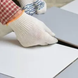 Układanie szarych i białych płytek podłogowych, widoczne dłonie w roboczych rękawicach, częściowo ułożona podłoga.