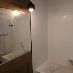 Wrocław: Fragment łazienki w trakcie wykończenia z białymi płytkami, drewnianą obudową lustra i wiszącą lampą. Wanna zabezpieczona folią ochronną.