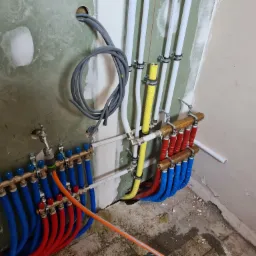 Rozdzielacze ogrzewania podłogowego z podłączonymi rurami w kolorach niebieskim i czerwonym, widoczne również białe i żółte rury instalacyjne, ściana w trakcie wykończenia.